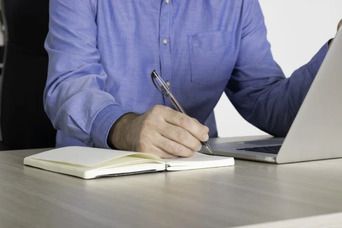 Businessman using laptop, taking notes at office desk - Close up. Man using computer and writing notes to notebook at office desk. Busy business man writing in an agenda on a desktop at office. cheap papers online