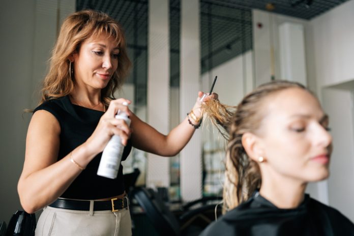 Lady client with long blonde hair getting stylish hairdo done at salon. Female hairdresser spraying hair setting liquid on blonde woman customer hair. Nicolas Krafft