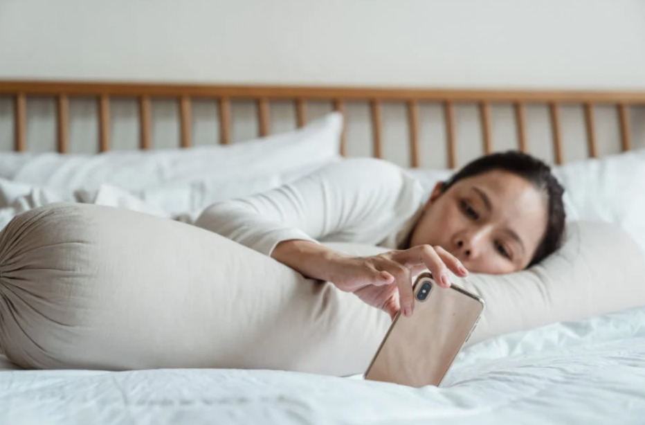 View of woman relaxed and lying in bed supported by a sausage pillow while passively interacting with her smartphone.
