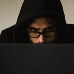 Close up view of suspicious-looking man’s face wearing a hood, slumped over working on a laptop.