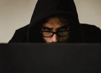 Close up view of suspicious-looking man’s face wearing a hood, slumped over working on a laptop.