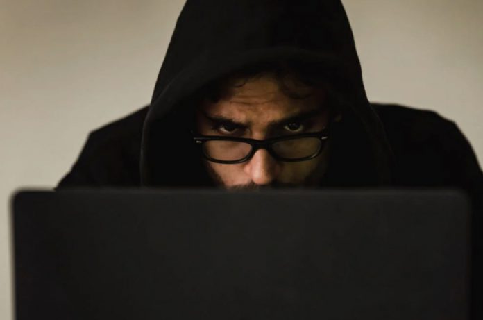 Close up view of suspicious-looking man’s face wearing a hood, slumped over working on a laptop.