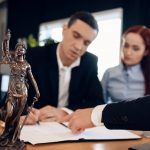 Statue of Themis holds scales of justice. In unfocused background, adult man signs documents.