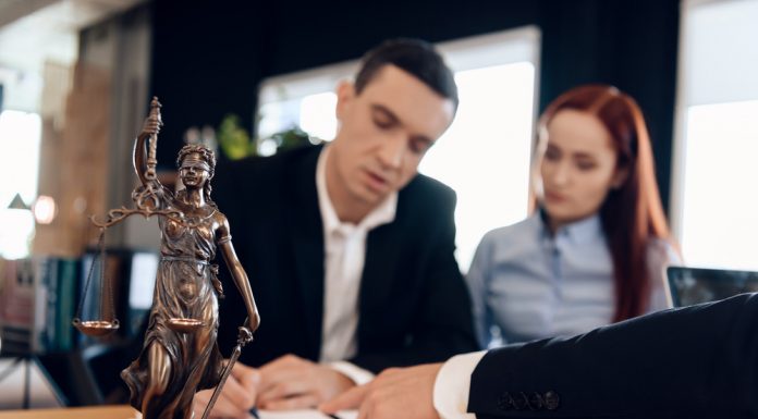Statue of Themis holds scales of justice. In unfocused background, adult man signs documents.