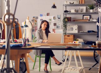 Female Fashion Designer In Studio Working On Sketches Or Documents At Desk With Laptop