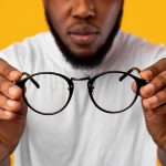African Man Holding Glasses Having Poor Eyesight, Yellow Background, Cropped