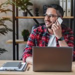 Portrait of smiling grey hair man with beard sitting front table with laptop, talking on phone