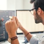 Precise Guidelines for Bitcoin Trading as a Beginner Investing. Close-up of young bearded trader in eyeglasses working with graph and financial reports on computer screen and holding Bitcoin in one hand while working his modern office.