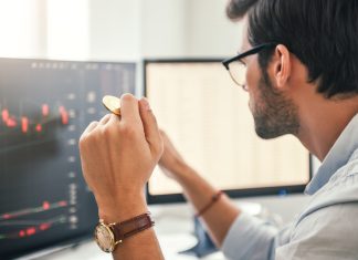 Investing. Close-up of young bearded trader in eyeglasses working with graph and financial reports on computer screen and holding Bitcoin in one hand while working his modern office.
