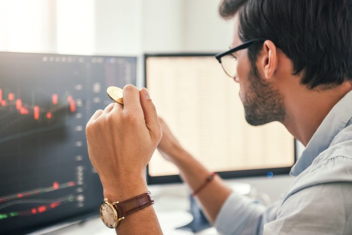 Investing. Close-up of young bearded trader in eyeglasses working with graph and financial reports on computer screen and holding Bitcoin in one hand while working his modern office.