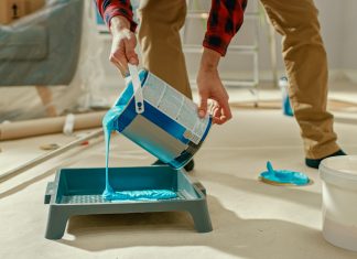 Common Mistakes That Can Happen When Renovating Your House! Young Man in Brown Jeans and Red Checked Shirt Picks Up a Can of Light Blue Paint and Pours it Into a Tray. Room Renovations at Home.