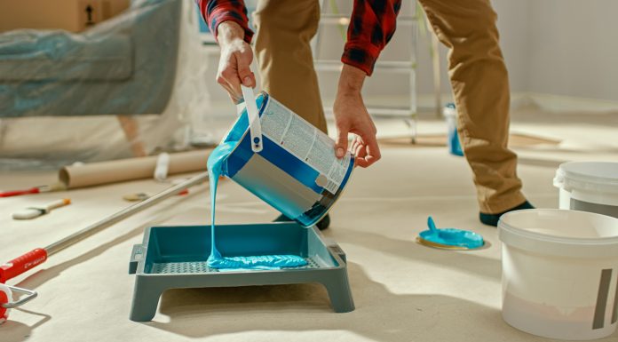 Young Man in Brown Jeans and Red Checked Shirt Picks Up a Can of Light Blue Paint and Pours it Into a Tray. Room Renovations at Home.