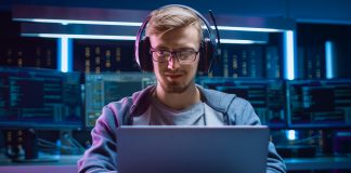 Learn Programming While Playing Computer Games Portrait of Software Developer / Hacker / Gamer Wearing Glasses and Headset Sitting at His Desk and Working / Playing on Laptop. In the Background Dark High Tech Environment with Multiple Displays.