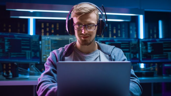 Portrait of Software Developer / Hacker / Gamer Wearing Glasses and Headset Sitting at His Desk and Working / Playing on Laptop. In the Background Dark High Tech Environment with Multiple Displays.