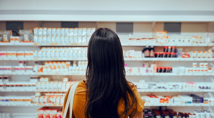Rear view of young woman with bag standing against shelf in pharmacy searching for medicine