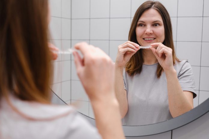 Close up of a woman in front of a mirror in the bathroom putting on invisible plastic teeth aligners or braces. Beautiful and healthy smile. Teeth whitening.