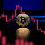 How Russia Invasion of Ukraine Can Affect the Crypto World A physical gold Bitcoin reflects light on a table with a laptop in the background. A chart is show which consists of red candlesticks in a downward trending pattern showing loss.