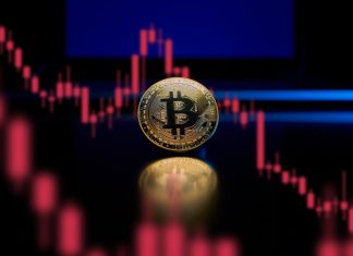 How Russia Invasion of Ukraine Can Affect the Crypto World A physical gold Bitcoin reflects light on a table with a laptop in the background. A chart is show which consists of red candlesticks in a downward trending pattern showing loss.