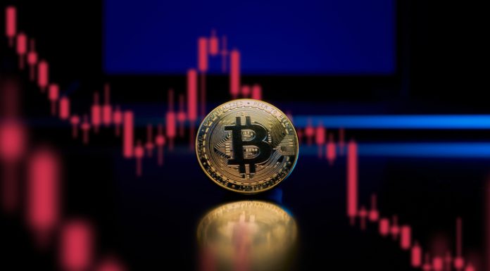 How Russia Invasion of Ukraine Can Affect the Crypto World A physical gold Bitcoin reflects light on a table with a laptop in the background. A chart is show which consists of red candlesticks in a downward trending pattern showing loss.