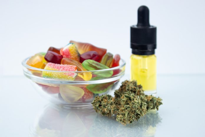 Assorted different gummy candies in a small glass bowl and a bottle of CBD canna oil, near in the foreground several buds of dried medical marijuana, reflected on the glass How CBD Gummies Can Help Relieve Anxiety