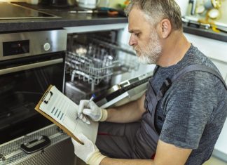 Male Technician Sitting Near Dishwasher Writing On Clipboard In Kitchen