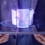 Close up of businessman hands holding tablet with creative digital blue cards hologram on dark background. Online casino, poker and digital gaming concept.
