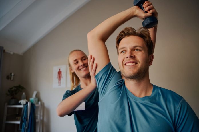 Portrait of handsome young smiling man exercising using dumbbells with female physiotherapist stock photo iStock-1271512414