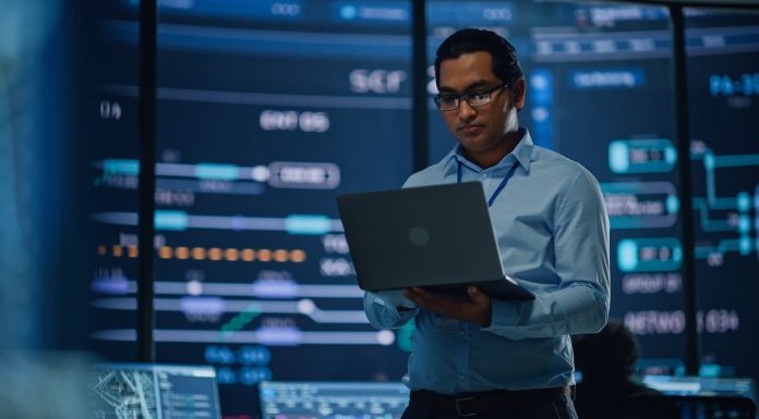 Young Multiethnic Male Government Employee Uses Laptop Computer in System Control Monitoring Center. In the Background His Coworkers at Their Workspaces with Many Displays Showing Technical Data.