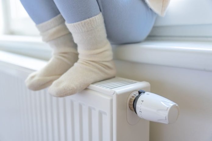Feet of little girl in warm white socks. Child sitting and warming up from the heating radiator. Focus on battery heating knob. Heating in an apartment, at home. Energy-Efficiency in the Spotlight
