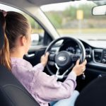 Woman driving car. Young driver. Testing vehicle in dealership or student training in school to get license. Hands on steering wheel. Behind back inside view of cockpit.