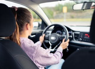 Woman driving car. Young driver. Testing vehicle in dealership or student training in school to get license. Hands on steering wheel. Behind back inside view of cockpit.