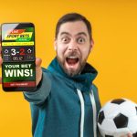 Happy excited football fan showing to the camera his smartphone with bookmaker's website application interface template. Focus is on the hand with phone. Isolated over yellow background.