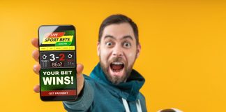 Happy excited football fan showing to the camera his smartphone with bookmaker's website application interface template. Focus is on the hand with phone. Isolated over yellow background.