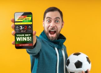 Happy excited football fan showing to the camera his smartphone with bookmaker's website application interface template. Focus is on the hand with phone. Isolated over yellow background.