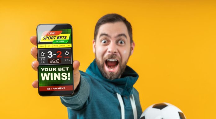 Live Gambling: A New Way to Play Happy excited football fan showing to the camera his smartphone with bookmaker's website application interface template. Focus is on the hand with phone. Isolated over yellow background.