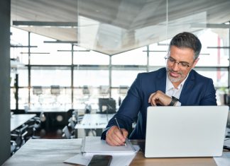 Mid aged business man working on laptop computer in office writing notes.