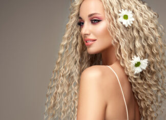 Portrait of young blonde haired beautiful model with flowers of chamomile in her long curly hairstyle. Soft smile on the attractive face.