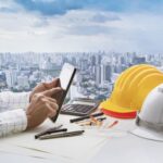 Hand of business man touching on computer tablet screen and safety helmet on working table against cities of high building for accommodation