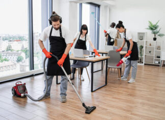 Team of young multicultural cleaners vacuums floor, wipes tables with gadgets, shelves, washes windows in spacious, bright, modern office. Professional cleaning company.