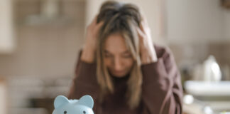 Overcoming A Scarcity Mindset A woman sits at a kitchen table with her head in her hands, looking at a piggy bank and a calculator.