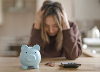 A woman sits at a kitchen table with her head in her hands, looking at a piggy bank and a calculator.