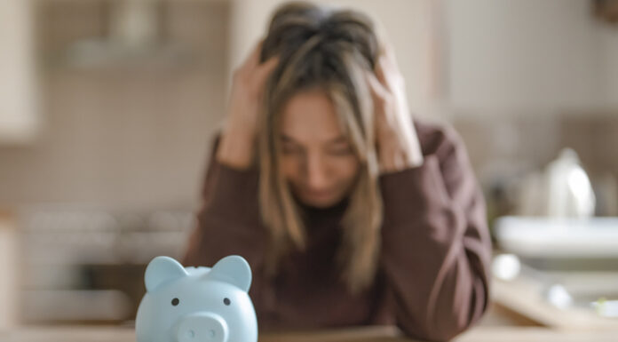 A woman sits at a kitchen table with her head in her hands, looking at a piggy bank and a calculator.