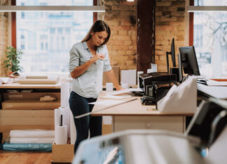 Portrait of charming woman in blue shirt looking at notes while standing near table with printer