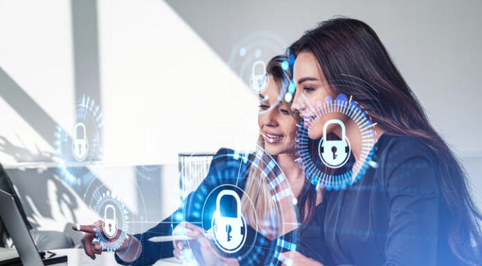 Businesswomen working together finger pointing at laptop on desk, smiling portraits. Glowing padlock icon with circuit hud. Cybersecurity and data protection.