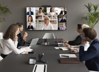 Employees speaking online to diverse freelancers on wall display and office workers at meeting table
