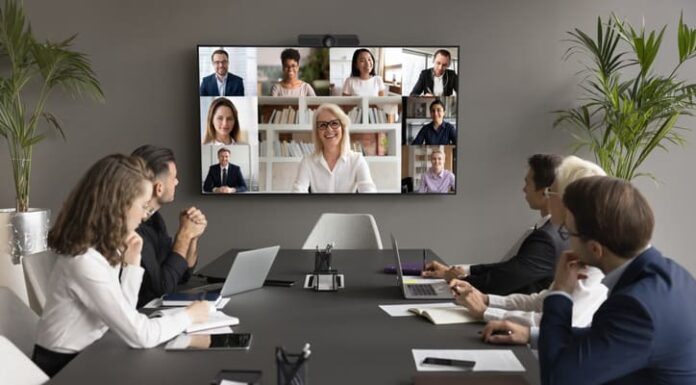 Employees speaking online to diverse freelancers on wall display and office workers at meeting table