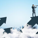 Businessman standing on abstract arrow and using telescope to look into the distance on city background.