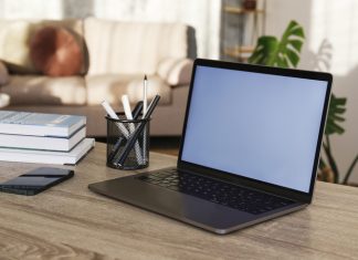 Blank screen laptop, cell phone and a cup on wooden table. Cozy lofty office with house plants full of natural sunlight - Business