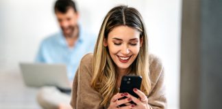 AsianMelodies Review: Check If It’s the Right Communication Platform For You Happy smiling young woman holding smartphone on floor at home using online entertainment mobile app/ AsianMelodies