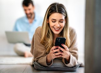 Happy smiling young woman holding smartphone on floor at home using online entertainment mobile app/ AsianMelodies